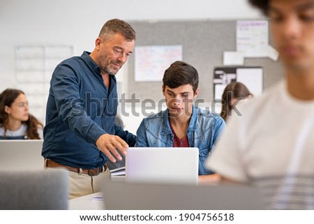Professor assisting college student with laptop in classroom during computer lesson. Teacher talking and explaining to guy. Mature man lecturer helping high school teen with laptop during lecture. Royalty-Free Stock Photo #1904756518