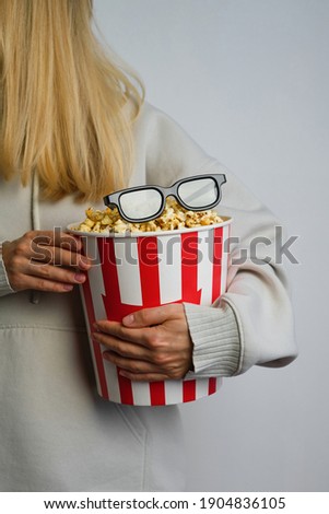 Popcorn paper bucket and 3D glasses in the hands of a young girl preparing to watch a movie. Showtime. Eating delicious unhealthy sweet snacks. Going to cinema for a new film. Rest and entertainment