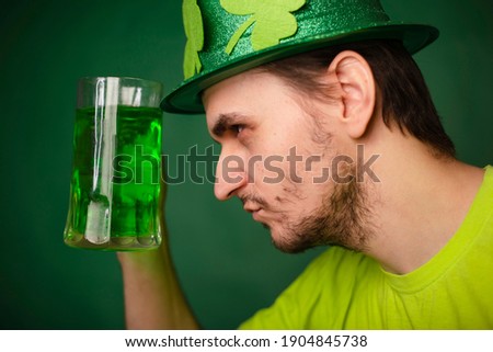 A man in a shamrock hat and green T-shirt drinks a large mug of green ale. Guy celebrates st patrick's day with a glass of beer.