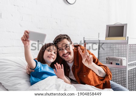 happy man showing victory gesture near daughter waving hand while taking selfie on smartphone, blurred foreground