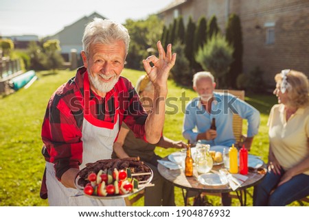 Senior man bringing delicious food on a tray while having an outdoor lunch with friends in the backyard of his house, showing an OK sign and smiling