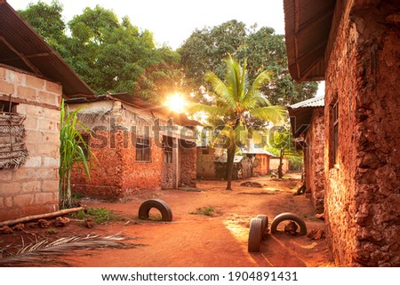 Scenic sunset over african village with palm. Street full of light, vibrant red soil and red color of buildings. Huts along the street. Traditional architecture, Tanzania countryside. Royalty-Free Stock Photo #1904891431