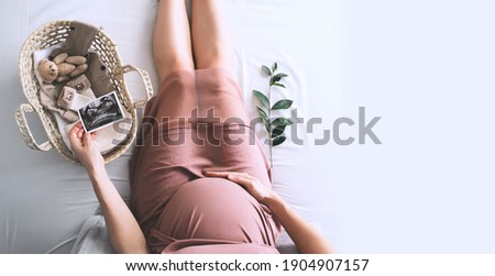 Pregnant woman in dress with ultrasound image. Mother with wicker basket of cute tiny stuff and teddy bear toy for newborn. Expectant mother waiting and preparing for baby birth during pregnancy. Royalty-Free Stock Photo #1904907157