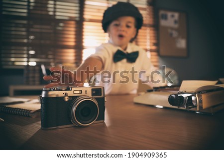 Cute little detective taking camera at table in office, focus on device