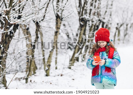 Little girl rolling up a funny video in smartphone. Happy child playing at winter forest use a smartphone look at the screen of a cell phone, watch cartoons.