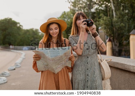 stylish young women traveling together in Europe dressed in spring trendy dresses and accessories smiling happy friends having fun taking photo on camera holding map
