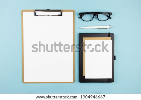 Modern office workplace with blank clipboard mockup, pen, glasses and envelope on blue background. Top view. Copy space. Flat lay.
