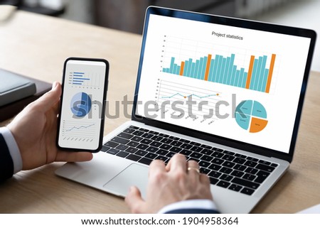 Close up young businessman analyzing project statistics working on computer and using smartphone application, developing marketing or sales strategy with modern technology gadgets in office. Royalty-Free Stock Photo #1904958364