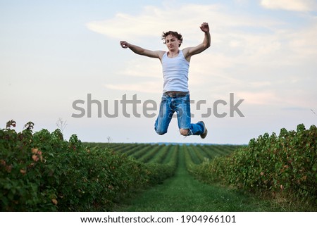 Young handsome man, wearing torn jeans and white t-shirt, jumping in the air on green field, smiling, laughing, having fun. Artistic education concept. Creative portrait outside. Freedom inspiration.
