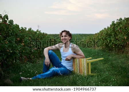 Young handsome man, wearing torn jeans and white t-shirt, sitting by yellow stool on green field, having fun. Artistic education concept. Creative portrait outside.
