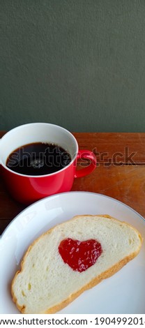 Heart-shaped strawberry jam on the bread. It was placed on a wooden table near the red coffee cup. The wall beside the table is gray.