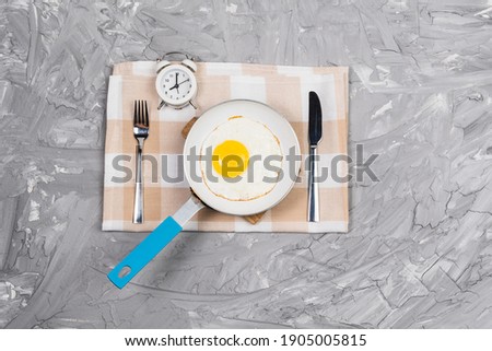 Alarm clock and frying pan fried egg with fork and knife. The concept of Breakfast