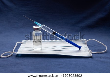 Covid-19 written vaccine bottle with liquid and syringe, standing on the protective mask on blue background