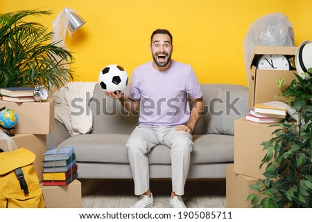 Shocked young owner man cheer up support favorite team with soccer ball sits in living room on sofa at home unpacking stuff rents flat isolated on yellow wall. Relocation moving in apartment concept