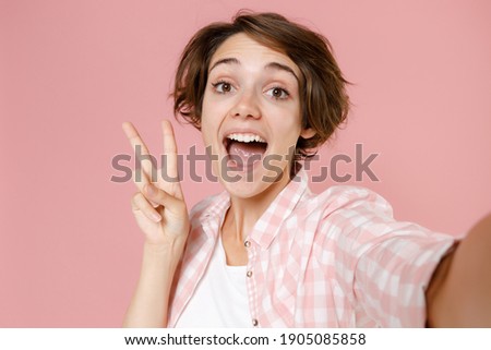 Close up of excited young brunette woman wearing casual checkered shirt standing doing selfie shot on mobile phone showing victory sign isolated on pastel pink colour background, studio portrait