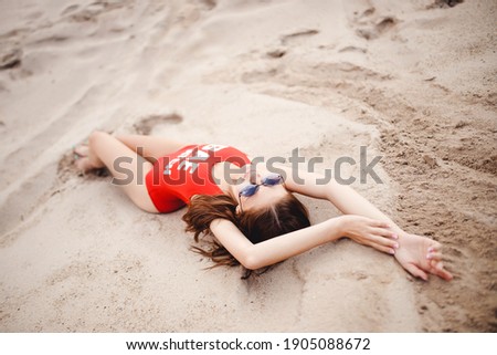 Relaxing on beach, young woman lies on sand in red swimsuit and sunglasses.