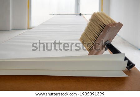 Wide paint brush for glue with wooden base for bristle,black handle,sheet of white wallpaper unfolded in length on table for pasting against renovating room with french window in backgdrop.Copy space Royalty-Free Stock Photo #1905091399