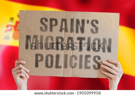 The phrase " Spain's migration policies " on a banner in men's hand with blurred Spanish flag on the background. Immigration. Travel. Explore. Move. Moving. Europe. EU. Rules. Law. Legislation