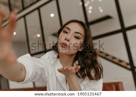 Brunette curly brown-eyed woman in white shirt takes selfie and blows kiss. Pretty lady in stylish outfit poses in living room.