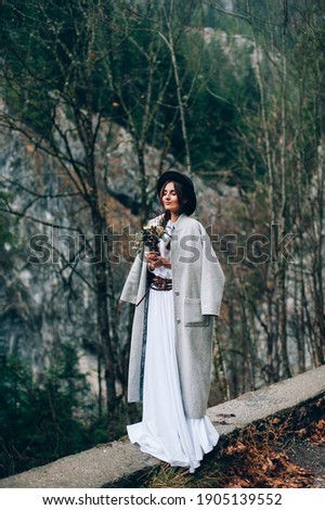 Stylish young girl. She is wearing a white dress and a coat with a hat on her head. In the hands of a gorgeous bouquet of flowers. During rainy weather in the mountains.