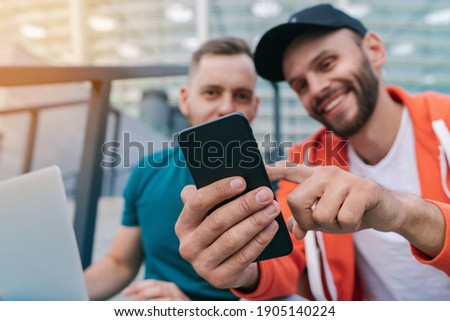 Two friends using mobile phone and laptop for betting during soccer play ready to celebrate victory of favourite team. Men using gambling application while sitting on stadium steps. Focus is on hand.