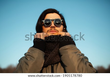 Close-up portrait of young guy with long hair wearing hipster round sunglasses and scarf. Blue sky on background.