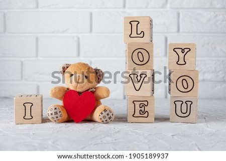 in the foreground are wooden cubes with letters lined with the phrase I love you, between the cubes sits a small teddy bear with a red heart, used as a background, tektur or postcard