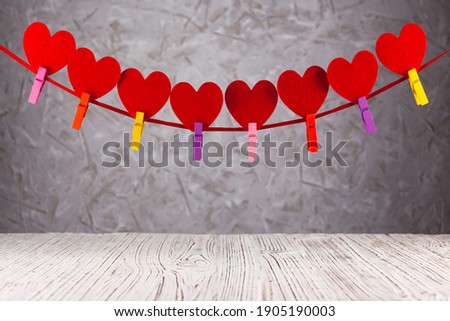 At the bottom of the frame is a wooden white surface, at the top hangs a garland with red hearts on the ribbon, attached with multi-colored clothespins, against the background textured background