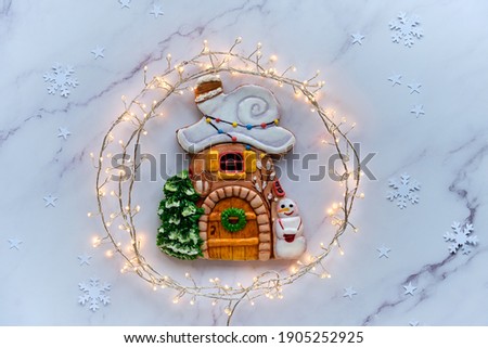 Decorated gingerbread house with creative glazing decor, snowman, Xmas tree. Christmas flat lay. Natural fir, stars and garland of festive lights. Top view, flat lay on white marble table.