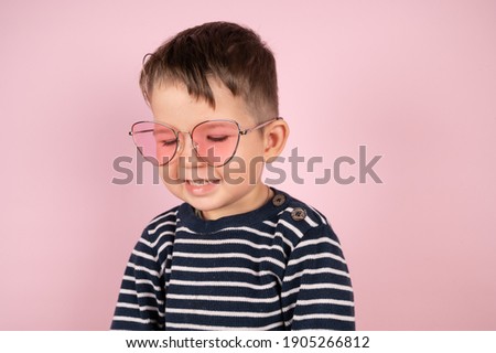 Cute toddler baby boy wearing pink sunglasses. Young guy boy in a striped sweater posing on a pink background, smiles and looks to the side , closeup.