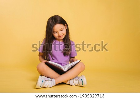 beautiful little girl in a purple t-shirt reads a book while sitting on a yellow background. Cute child learns lessons. Photo in studio