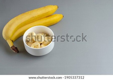 Two bananas and slices in a white plate on a gray background. Trending color of year 2021 Illuminating and Ultimate gray. Side view minimal still life with copy space.