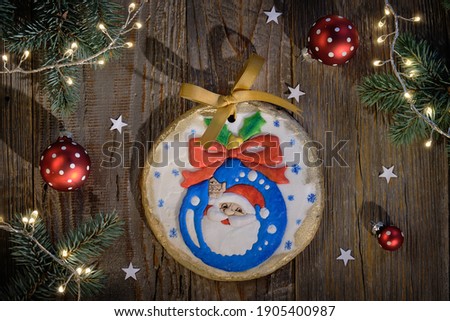 Ornate gingerbread decorated with Santa smiling from Xmas bauble. two cartoon cow characters. Christmas flat lay with fir twigs, red baubles, light garland. Top view, dark rustic wood background.