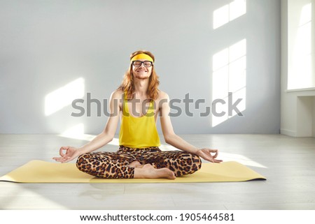 Happy funny relaxed smiling skinny man with hippie style long hair in nerd glasses, yellow retro headband, tank top and leopard print leggings meditating eyes closed in lotus yoga pose on exercise mat