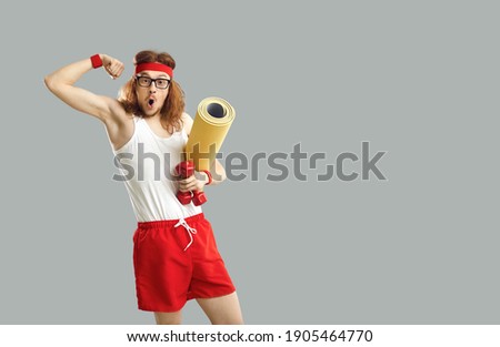 Funny happy man in fitness wear standing against gray free space for text studio background, holding sports workout mat and gym dumbbells and showing biceps muscles surprised at how big they've grown