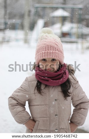 Little girl in winter clothes, a scarf and a hat with hands in pockets on the background of a snow-covered playground
