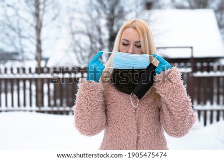 double mask. A blonde girl, outdoors in winter, wears a blue medical mask and holds the second black mask in her hand, in blue disposable gloves.