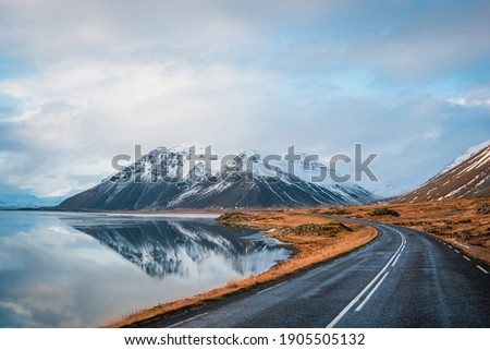 Panoramic winter photo of road leading along coast of lake to volcanic mountains. High rocky peaks covered with snow layer mirroring on water surface. Driver's point of view on Ring road, Iceland.
