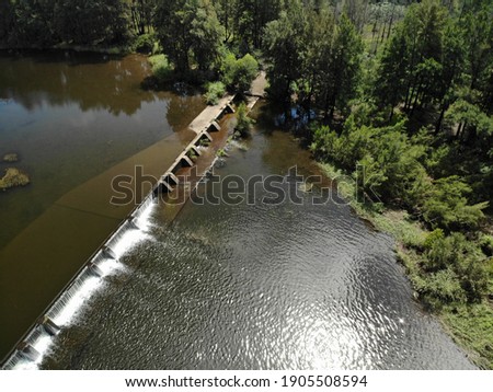 Aerial view of murky brown water gushing over the Nepean River Weir on a sunny summer day, sun reflecting off water, sparkling, river weeds, lush green trees. Penrith NSW, Australia