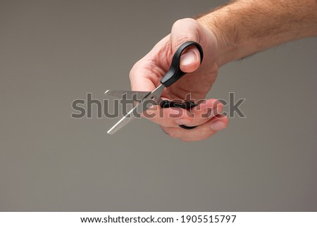 Caucasian male hand holding a small craft scissors with black plastic handles isolated on gray background.