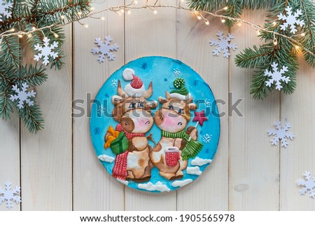 Decorated gingerbread with two cartoon cow characters. Christmas flat lay with fir twigs and snowflakes. Off white wooden planks, top view of rustic wooden table.