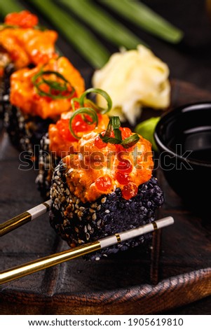 Japanese food. Dark and moody photo. Macro view of sushi roll. Black rice roll with fish. Chopsticks holding black sushi roll. Rolls on dark wooden board.