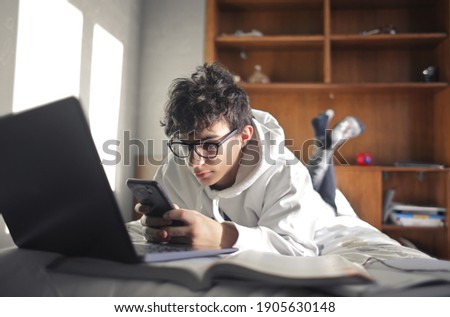 young boy studies lying on the bed using computer and smartphone Royalty-Free Stock Photo #1905630148