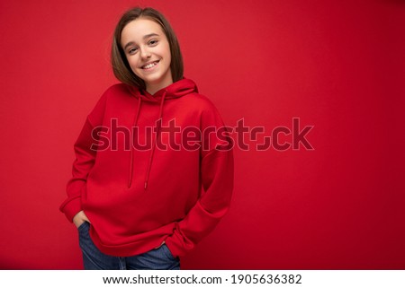 Beautiful positive smiling brunet little lady wearing stylish red hoodie standing isolated over red background wall looking at camera