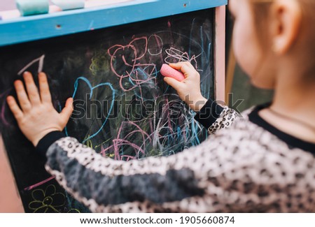Little preschool girl, child draws with hand with colored chalk on a black wooden board, doodle picture. Drawing lesson at school.