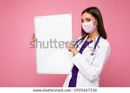 Beautiful brunette female nurse in protective face mask and white medical coat holding an empty magnetic board isolated on pink background