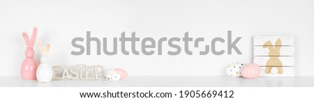 Easter decor on a white shelf. Rustic wood signs, eggs and modern glass bunnies against a white wall banner. Copy space.
