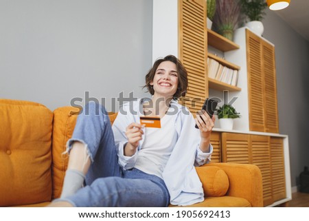 Smiling young woman in casual clothes sit on couch spending time in living room at home. Rest relax good mood leisure lifestyle concept. Using mobile phone, hold credit bank card doing online shopping