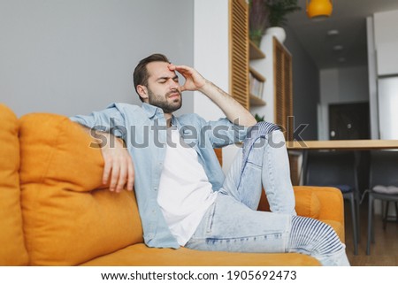 Exhausted tired young bearded man wearing casual white t-shirt blue shirt keeping eyes closed put hand on head having headache sitting on couch resting relaxing spending time in living room at home