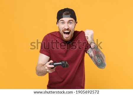 Screaming gambling young guy in casual t-shirt black cap play game with console joystick clenching fist isolated on yellow background studio portrait. Mockup copy space. Tattoo translate life is fight